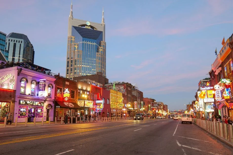 Bustling downtown street at dusk with neon signs along both sides and a tall glass skyscraper in the background.