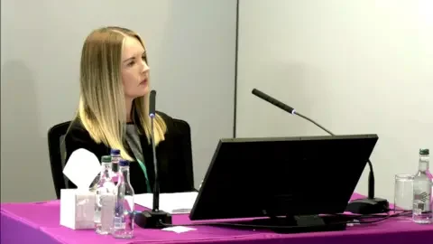 Lampard Inquiry Service manager Chloe Cawston sits in front of a table at the Lampard Inquiry. She is wearing a black jacket and a grey top underneath it. There are two microphones pointing torwards her and a laptop in front of her, to each side there are bottles of still and sparkling water and a box of tissues, the walls are white, the tablecloth is purple and she has long blonde hair.