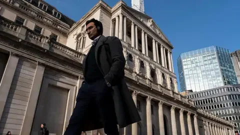 Mike Kemp/In Pictures via Getty Images A man in a long dark coat is striding past the Bank of England building on a sunny day.