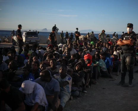 Police officers stand guard over migrants seated on the ground after crossing into Ceuta, Spain