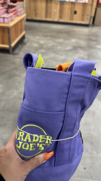 Purple Trader Joe’s Halloween mini tote with green and yellow arm straps, lying on a basket with orange juice.
